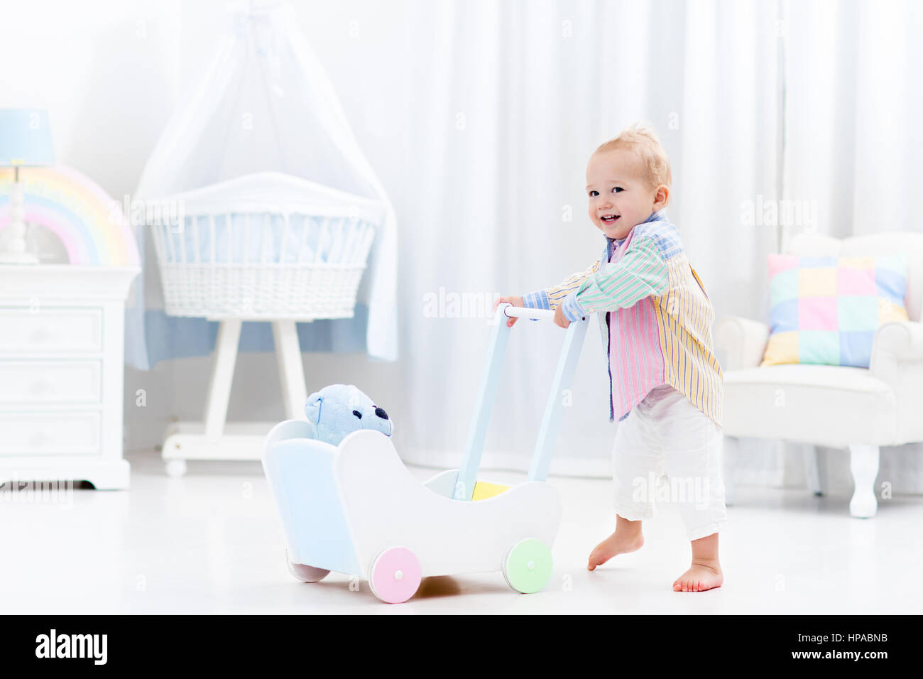 Baby boy learning to walk with wooden push walker in white bedroom with pastel rainbow color toys. Aid toy for child first steps. Toddler kid walking  Stock Photo