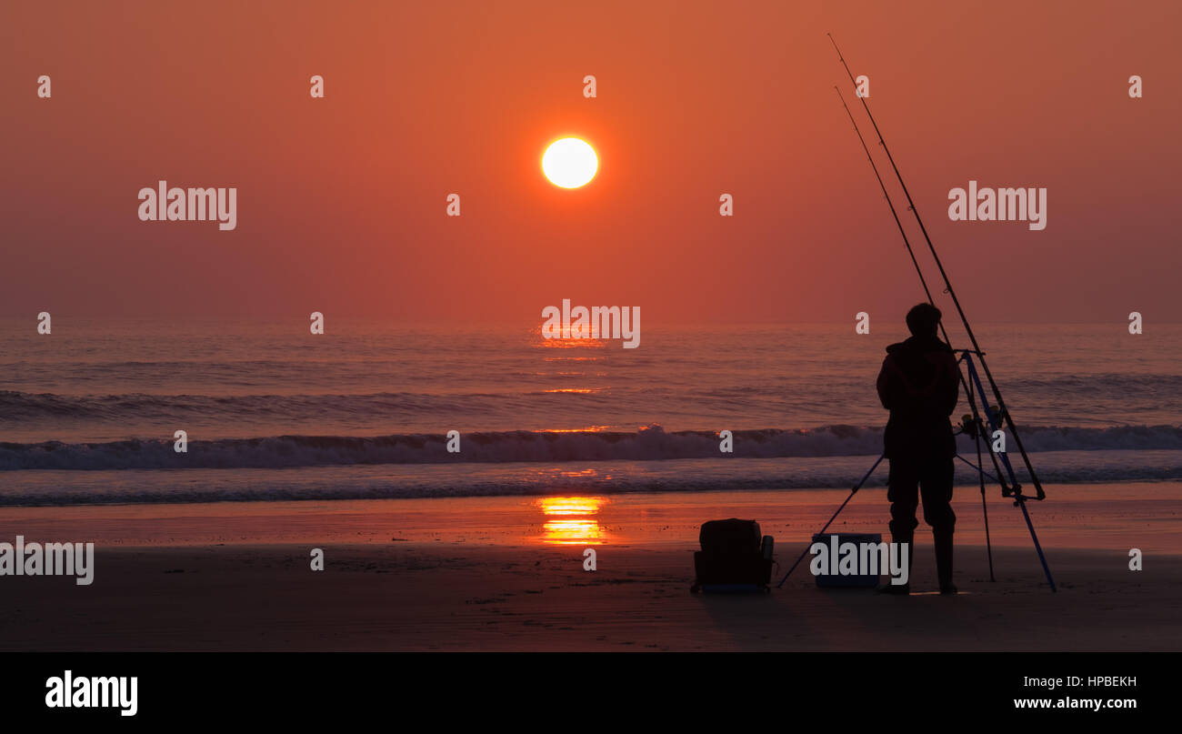 Catching A Sunset on The Beach At Trecco Bay Near Porthcawl In Wales. Alone Fisherman hoping for a catch as The Sunsets Stock Photo