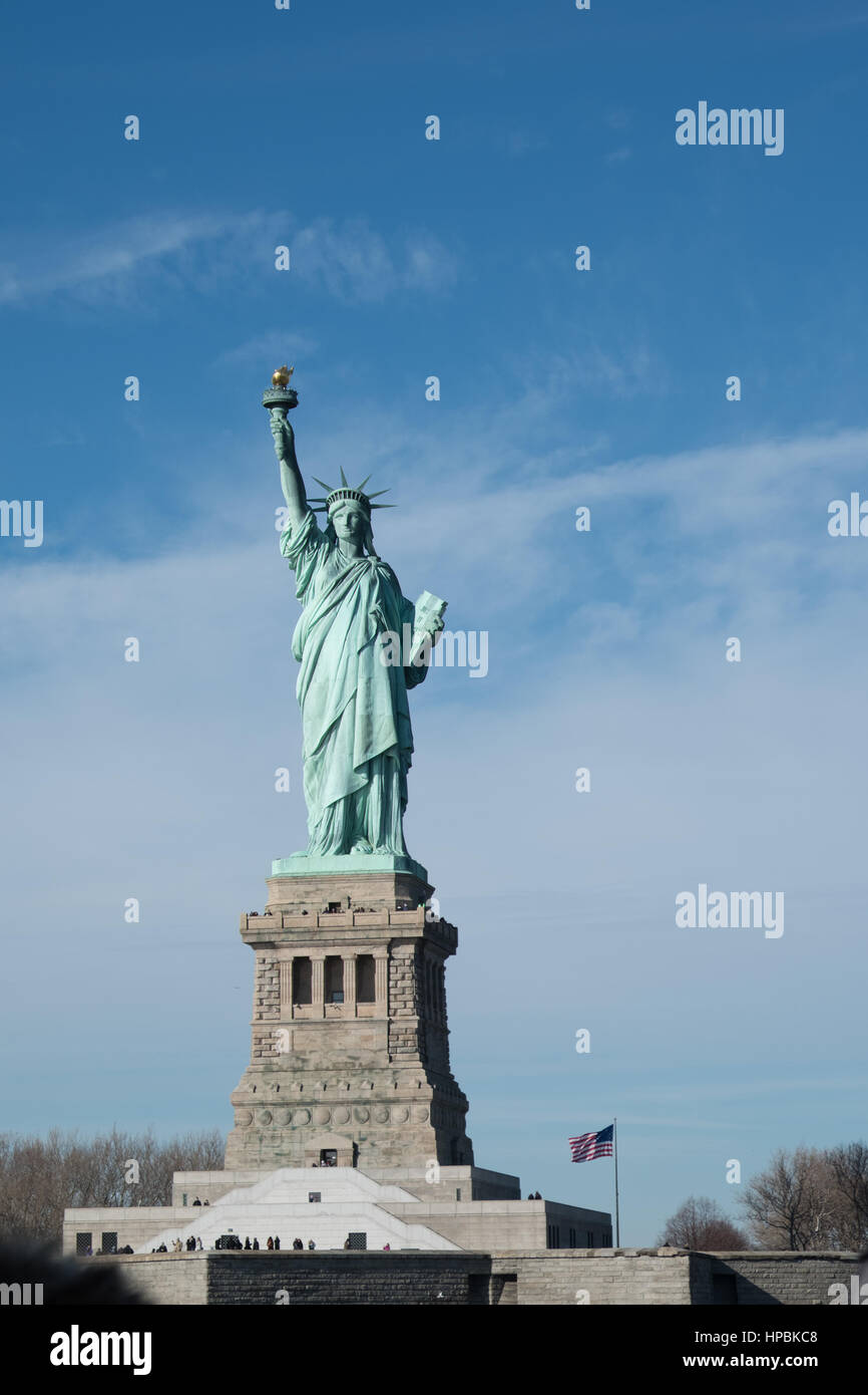 Statue of Liberty on Liberty Island, American Flag on a flagpole waving in background. Statue of Liberty on a bright sunny day with blue sky and scatt Stock Photo