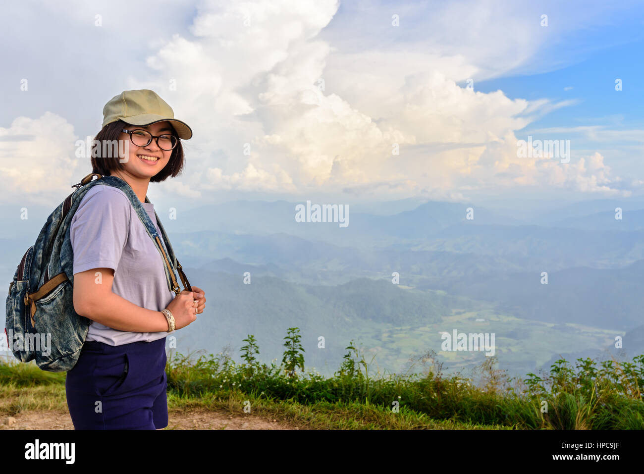 Teens girl hiker wear a cap and glasses with backpack is standing and smiling happily on high mountain at scenic point of Phu Chi Fa Forest Park Stock Photo