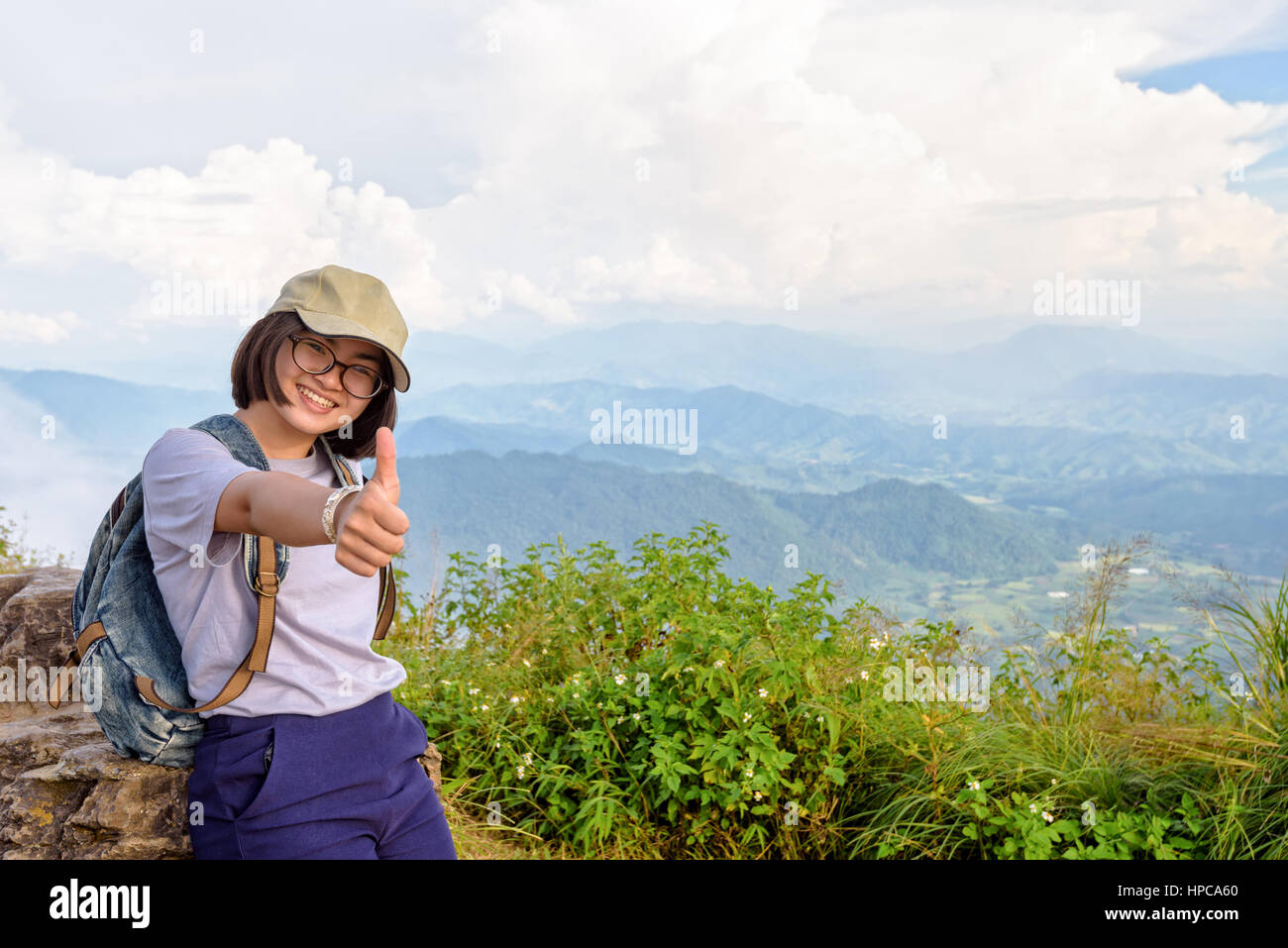 Tourist teens girl hiker wear cap and glasses with backpack poses thumb up smiling happily on high mountain at scenic point of Phu Chi Fa Forest Park  Stock Photo