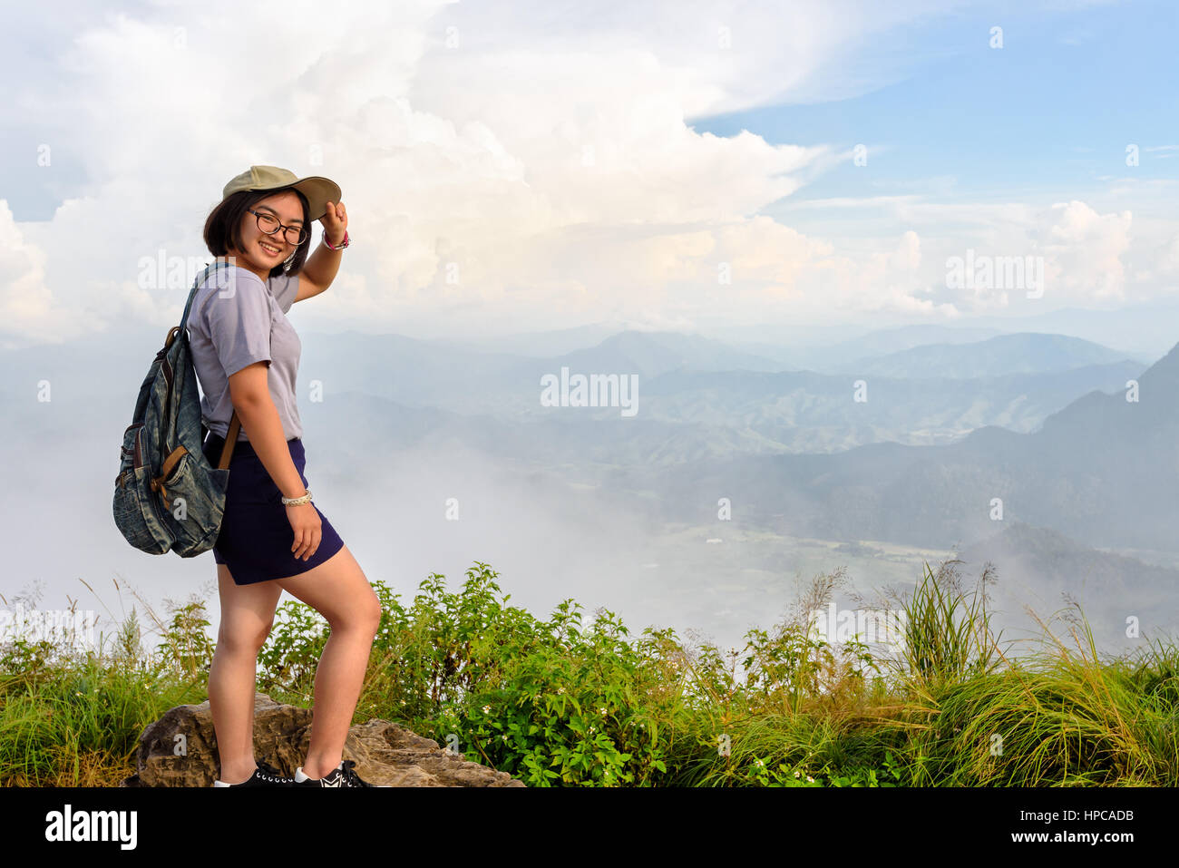 Tourist teens girl hiker with backpack cap and glasses is standing smile and poses happily at high mountain on sky and fog background at scenic point  Stock Photo