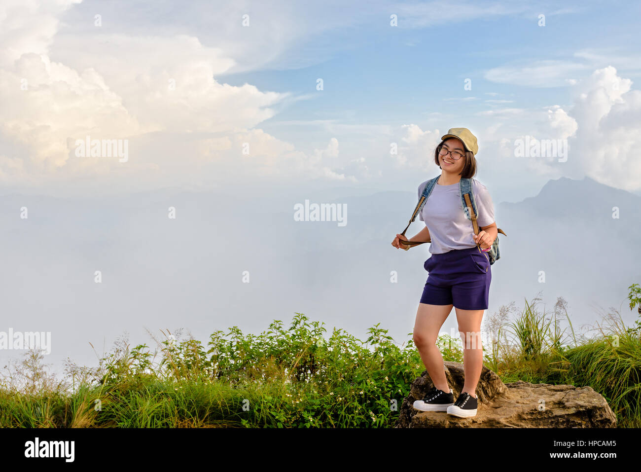 Tourist teens girl hiker with backpack cap and glasses is standing smile and poses happily at high mountain on sky and fog background at scenic point  Stock Photo