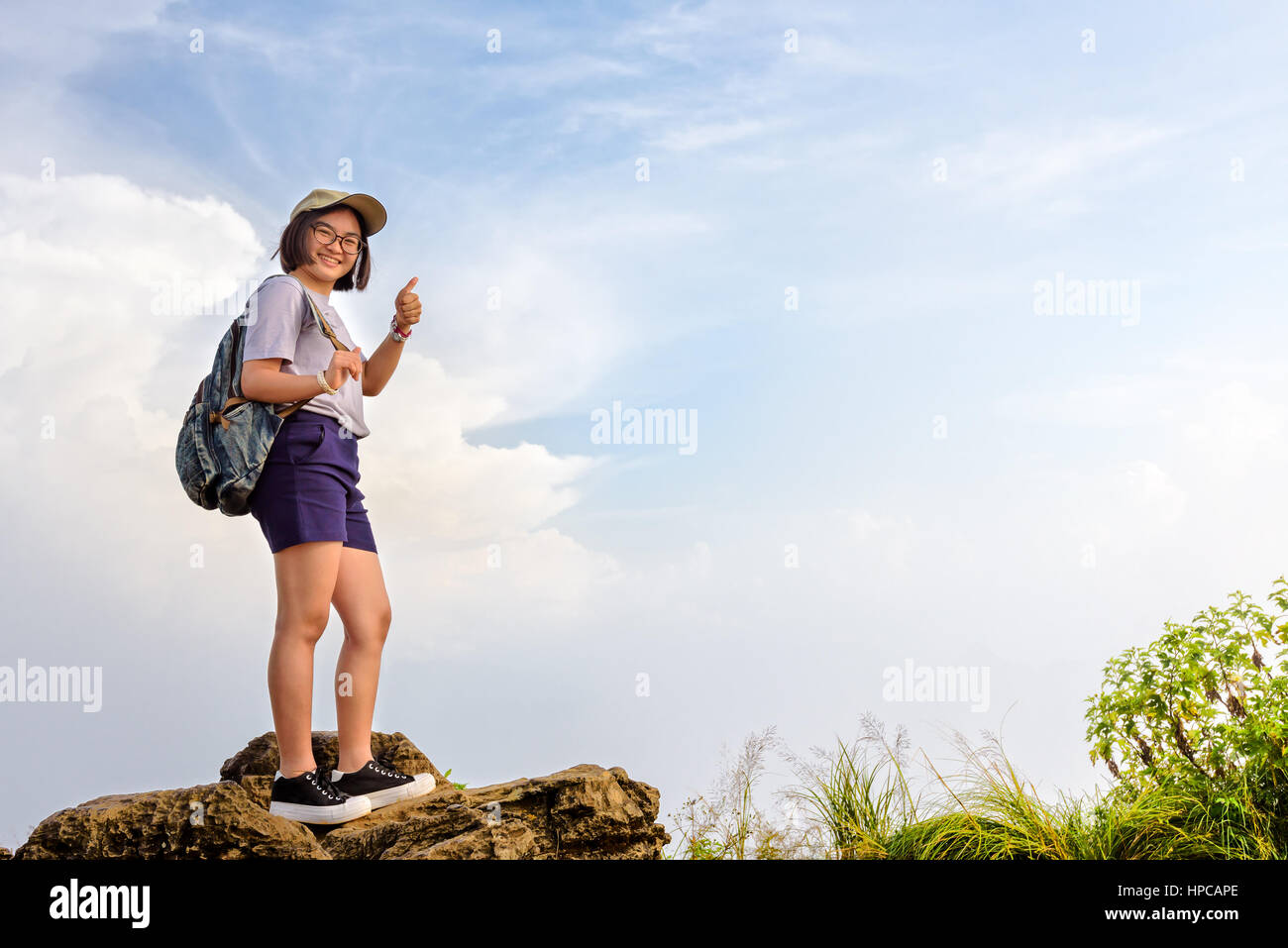 Happy hiker asian cute teens girl with backpack cap and glasses standing smiling poses thumb up on mountain and sky background at Phu Chi Fa Stock Photo