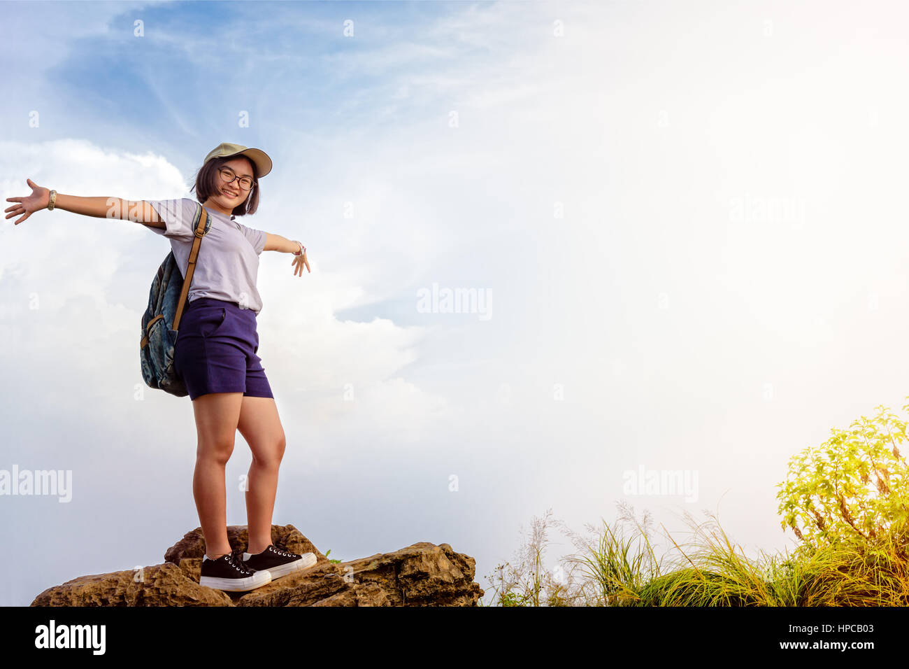 Happy hiker asian cute teens girl with backpack cap and glasses standing smiling poses open arms on mountain and sky background at Phu Chi Fa Forest Stock Photo