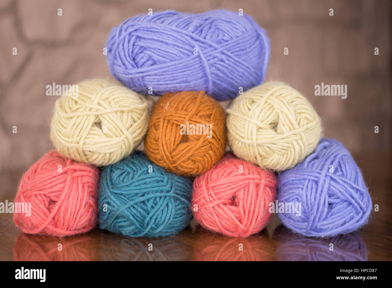 Eight skeins of lavender, gold, turquoise, cream and pink yarn stacked in a pyramid on a wood table. A stone wall is in background. Stock Photo