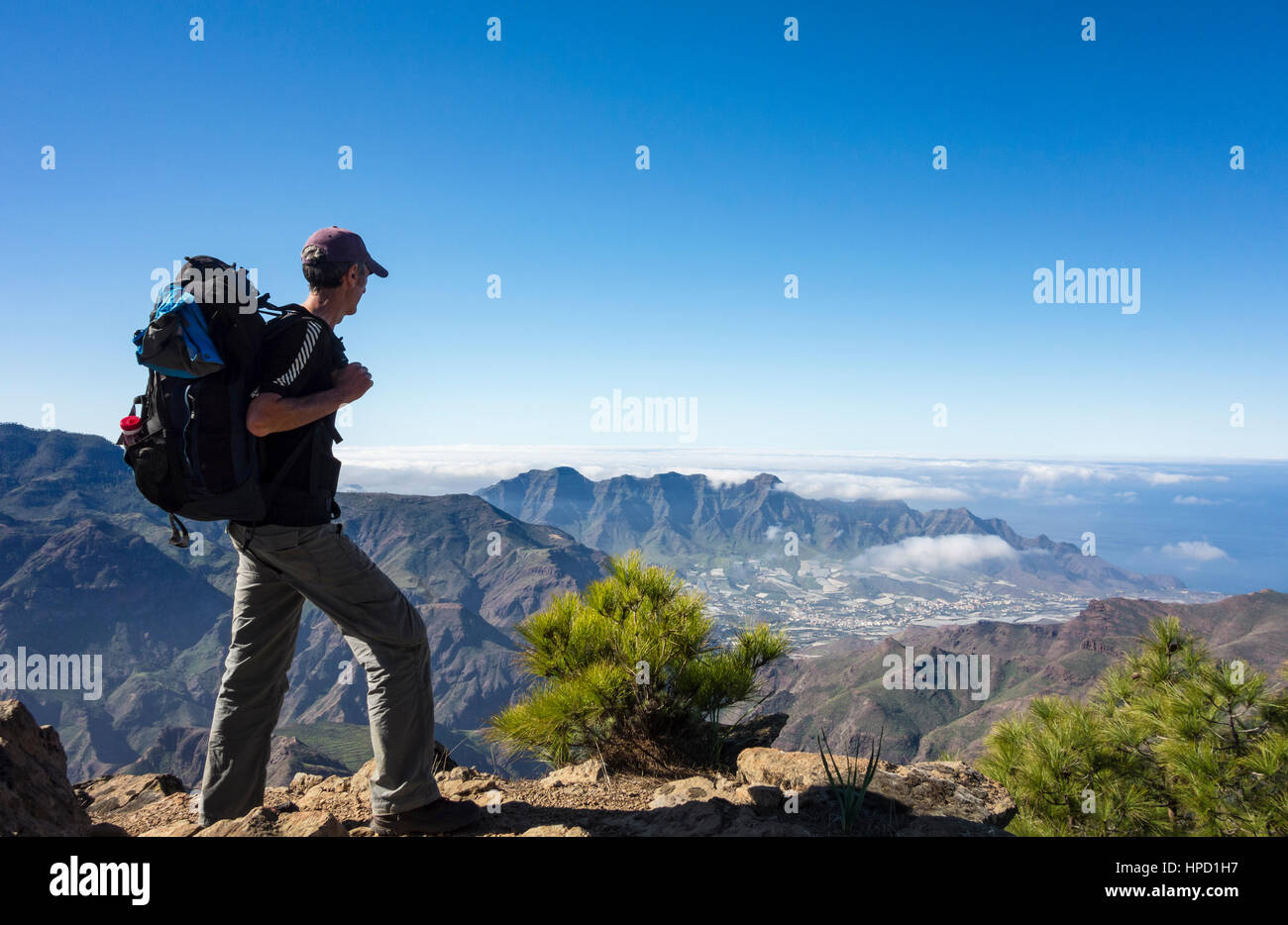 Mature male hiker on Alta Vista mountain on Gran Canaria with La Aldea de San Nicolas village in distance. Canary Islands. Spain Stock Photo