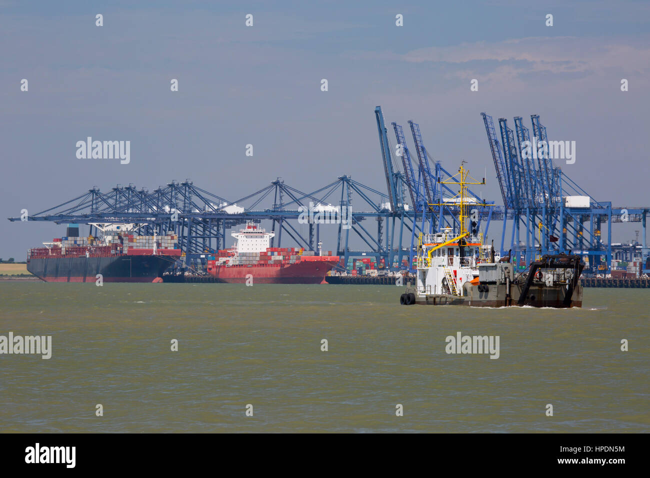 A line of container ships and cargo freighters berthed at Felixstowe container port, being unloaded or loaded, with a tug in the foreground. Stock Photo
