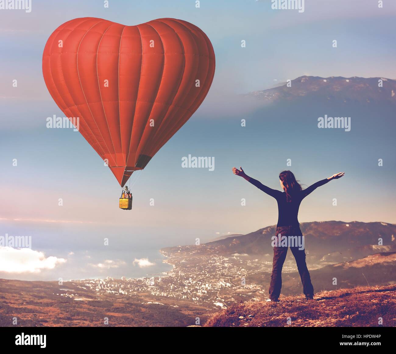 Girl with rised hands on the top of mountain looking at big red air balloon heart shape flying in blue sky. Romantic on Valentine's Day. Sport and rec Stock Photo