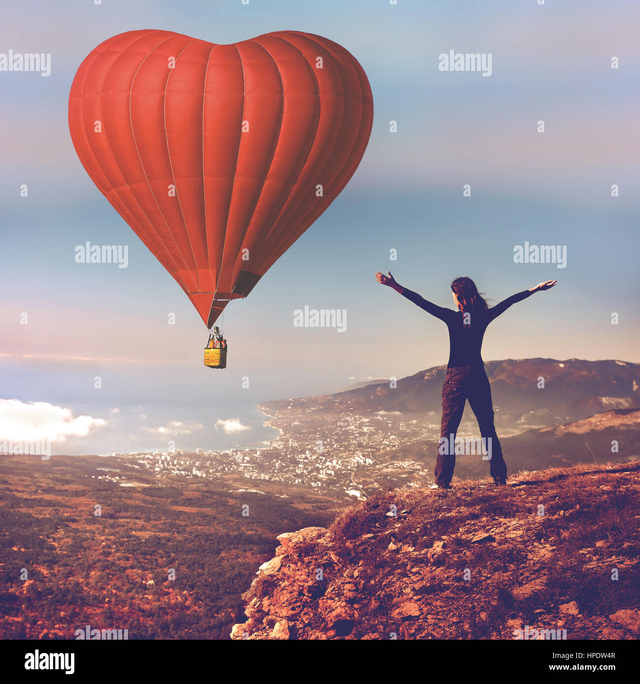 Girl with hands up stand on top of mountain looking at big red air balloon heart shape flying in blue sky. Romantic trip on Valentine's Day. Sports, t Stock Photo