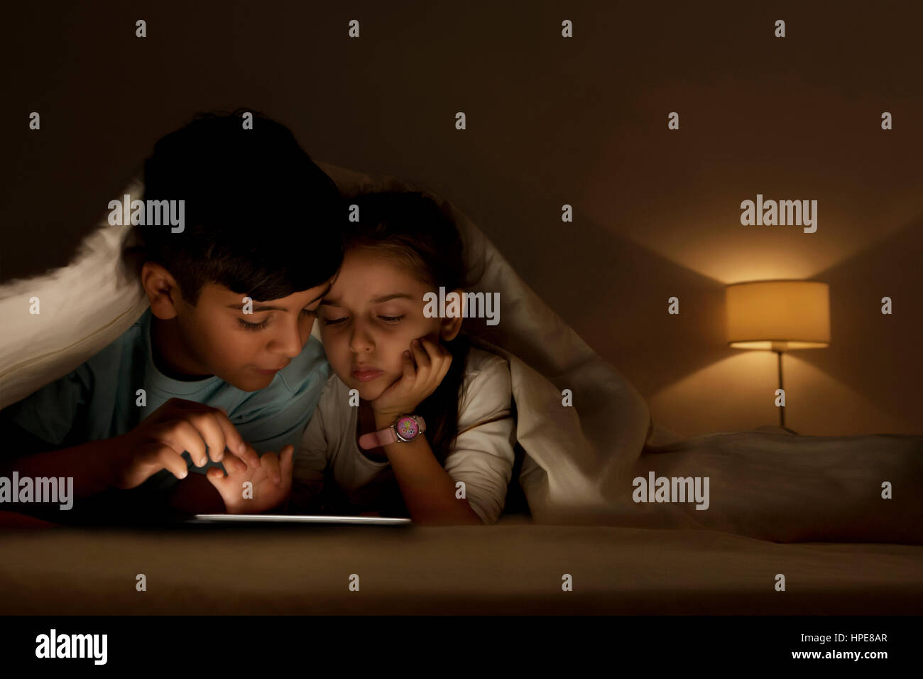 Brother and sister using digital tablet lying in bed under the covers Stock Photo