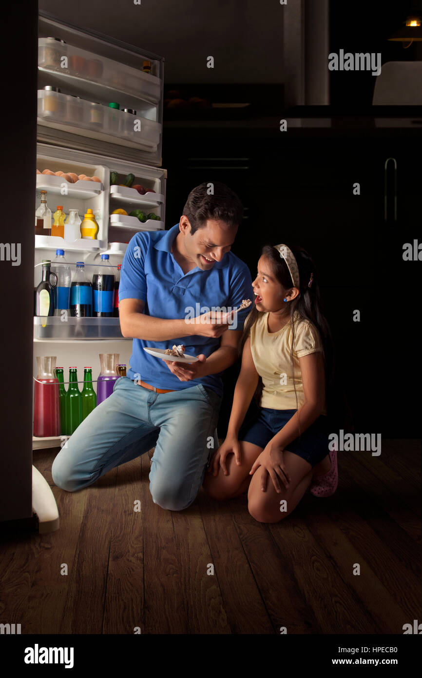 Father and daughter kneeling down in front of open refrigerator and eating cake Stock Photo