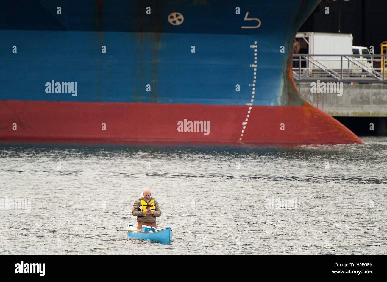 Drew Heifetz paddling his canoe out to check his crab pots by a cargo ship docked at the Squamish Terminal pier.  Squamish BC, Canada. Stock Photo