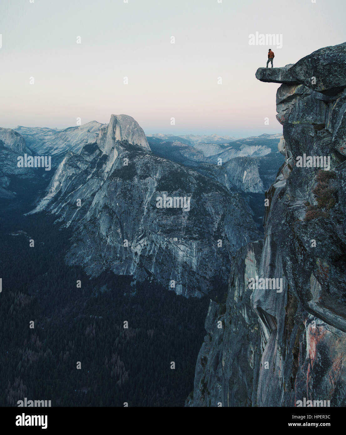 A fearless hiker is standing on an overhanging rock enjoying the view towards famous Half Dome at Glacier Point overlook in twilight, Yosemite NP, USA Stock Photo