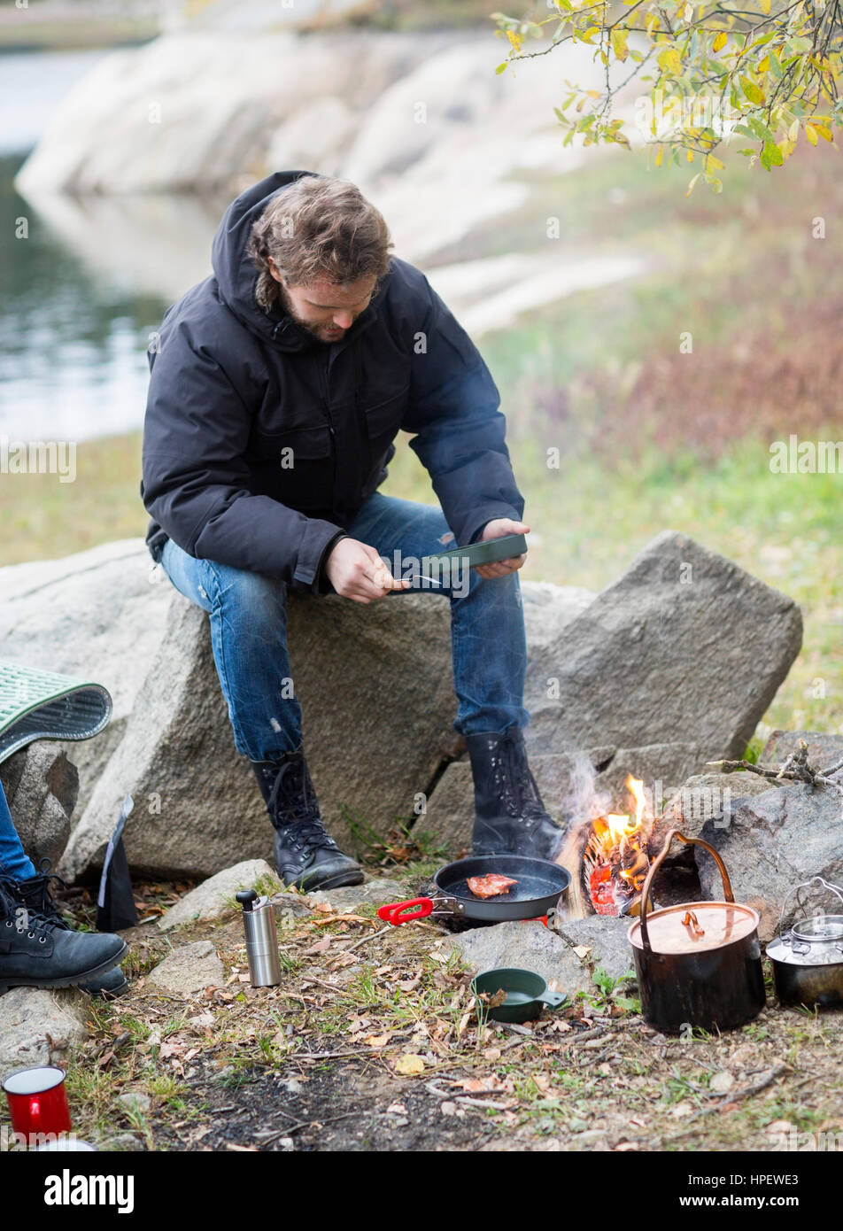 Full length of young man having food while sitting on rock at campsite ...