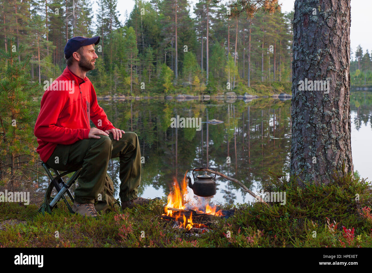 Hiker boiling water in tin kettle over camp fire while camping along lake Stock Photo