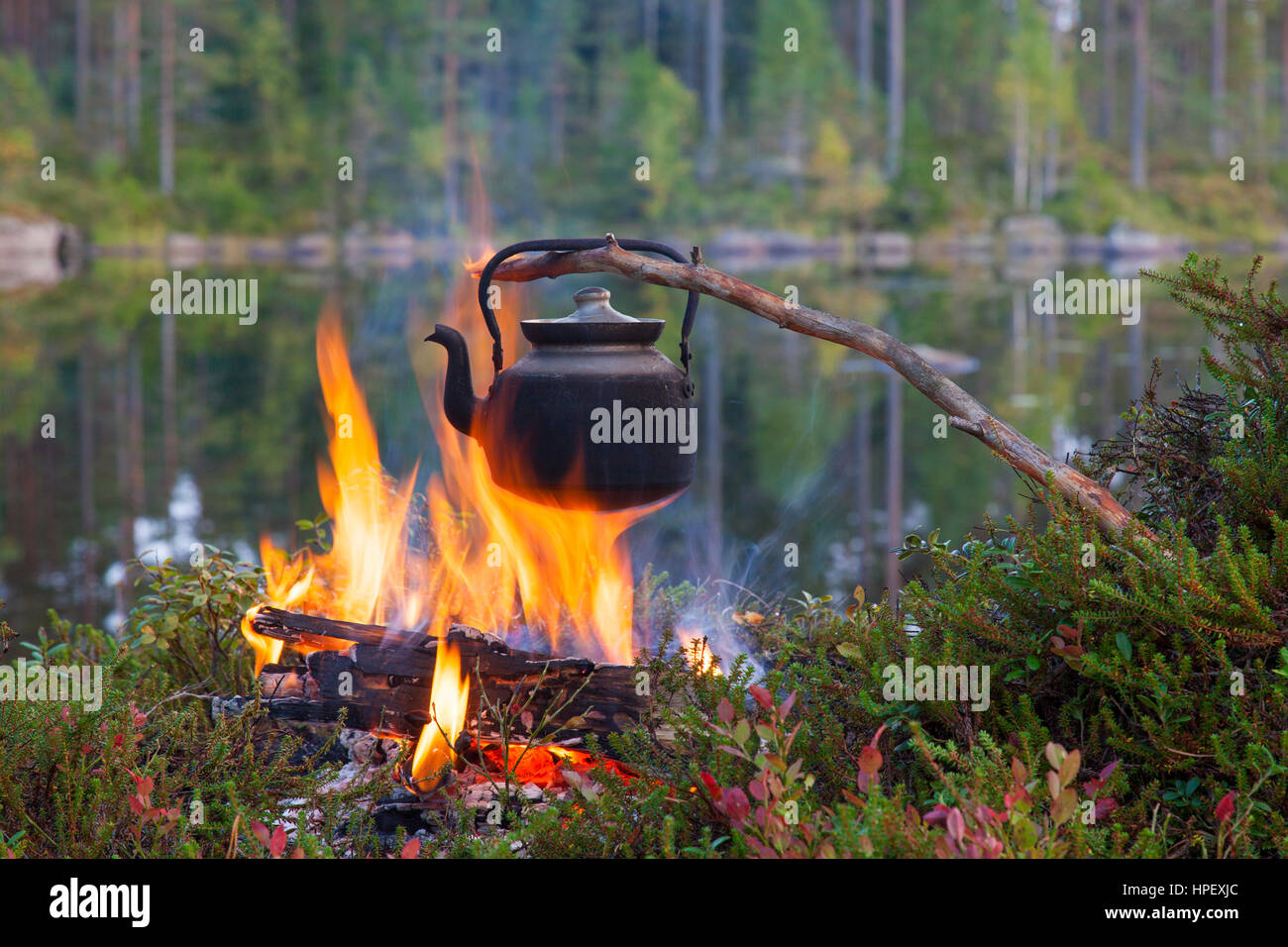 Blackened tin kettle boiling water over flames from campfire during hike along lake Stock Photo