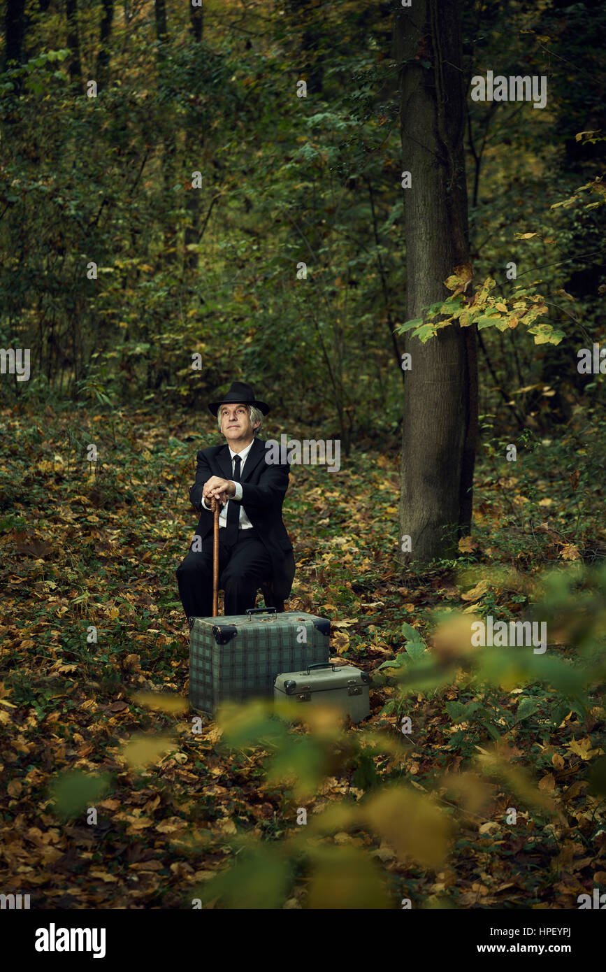 Man sitting in the forest with hat and stick and two suitcases Stock Photo