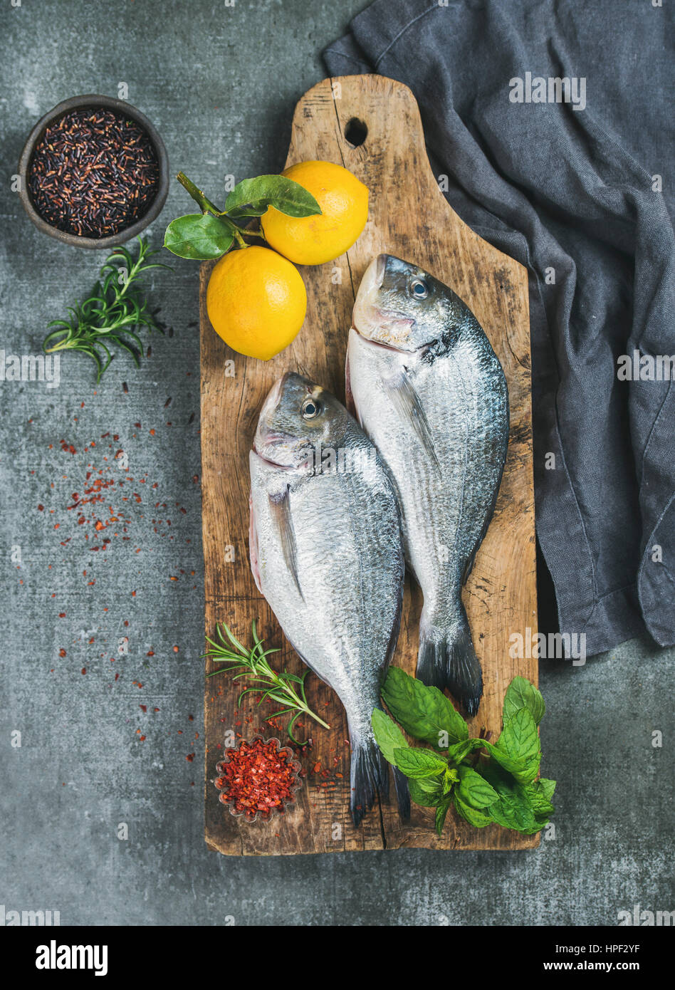 Fresh uncooked sea bream or dorado fish with lemon, herbs and spices in bowls on rustic wooden board over grey concrete background, top view. Healthy, Stock Photo