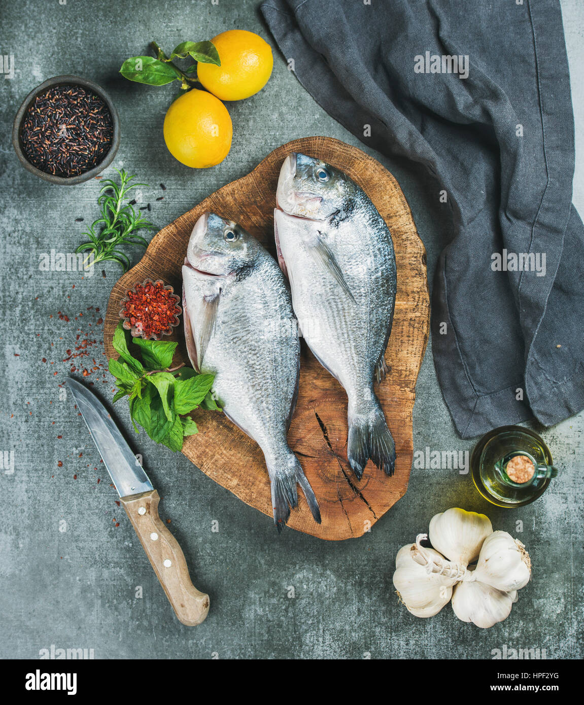 Fresh uncooked sea bream or dorado fish with lemon, herbs, olive oil, garlic and spices in bowls on rustic wooden board over grey concrete background, Stock Photo