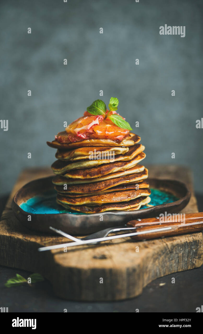 Homemade pancakes with honey, bloody orange slices and mint leaves for breakfast on blue ceramic plate over rustic wooden board, grey plywood wall at Stock Photo