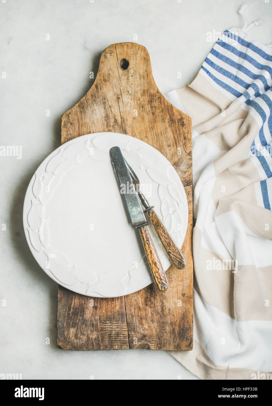 Mediterranean style table setting. Rustic wooden chopping board, white ceramic dinner plate, vintage cutlery and striped blue and white towel over gre Stock Photo