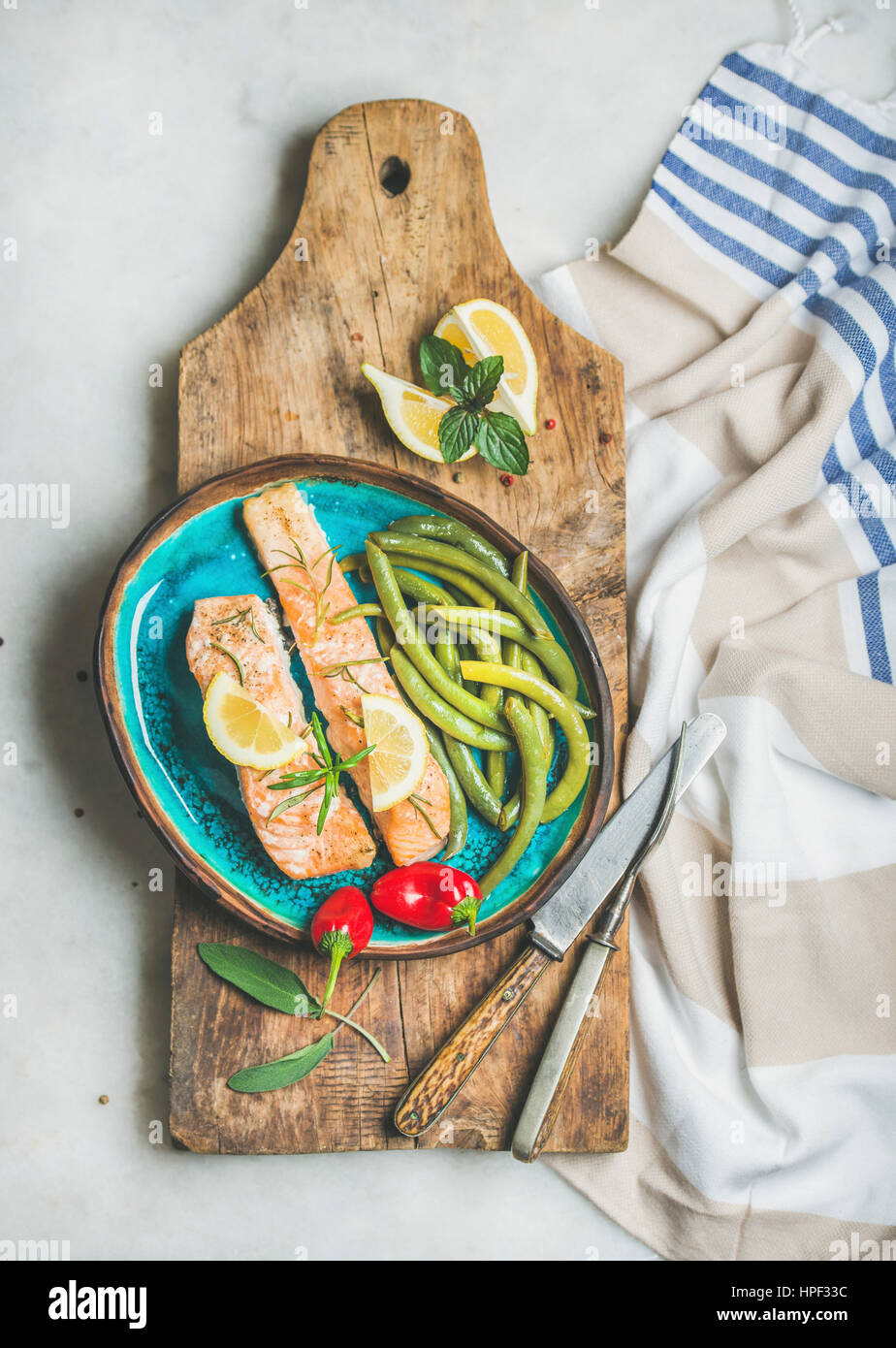 Oven roasted salmon fillet with lemon, rosemary, chilli pepper, poached green beans in blue ceramic plate on wooden board over grey marble background, Stock Photo