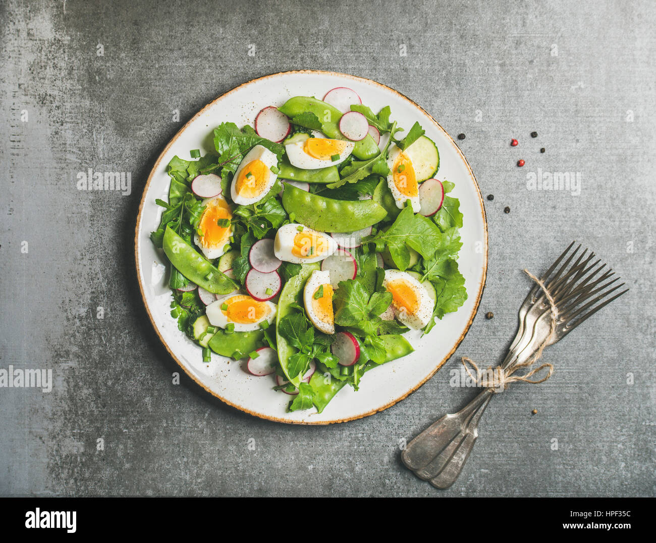 Healthy spring green salad with radish, boiled egg, arugula, green pea and mint in white plate over grey concrete background, top view Stock Photo