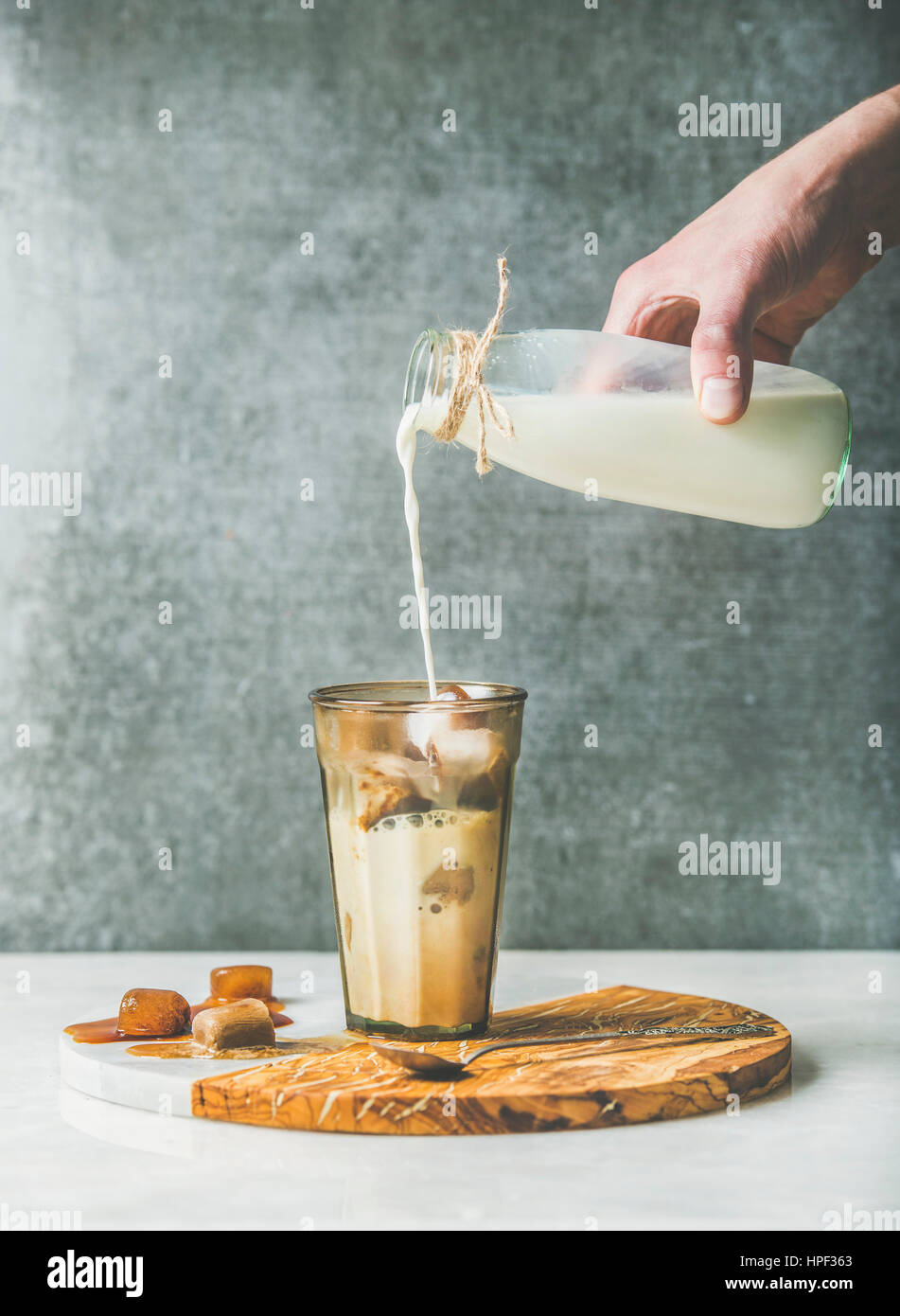 Man's hand pouring milk to Iced caramel latte coffee cocktail with frozen coffee ice cubes in glass on serving wood and marble board over grey table, Stock Photo