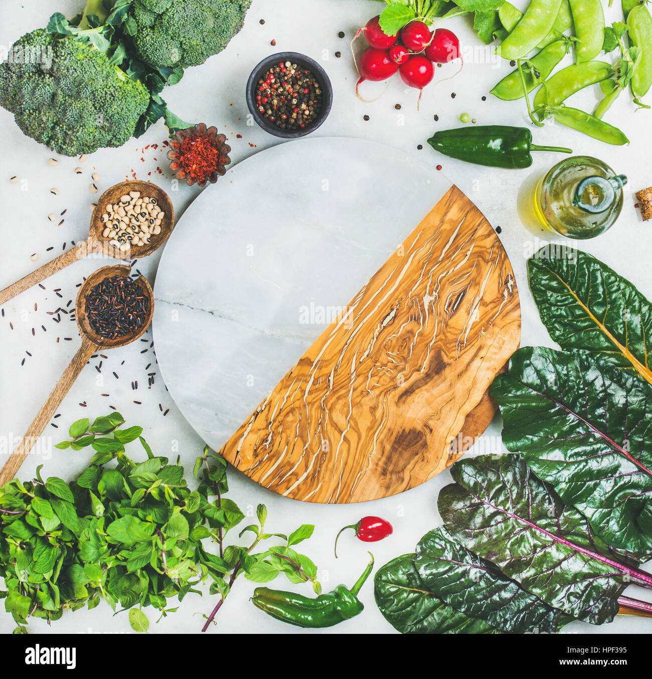 Fresh raw greens, vegetables, olive oil and grains over light grey marble kitchen countertop, round board in center, top view, copy space. Healthy, cl Stock Photo