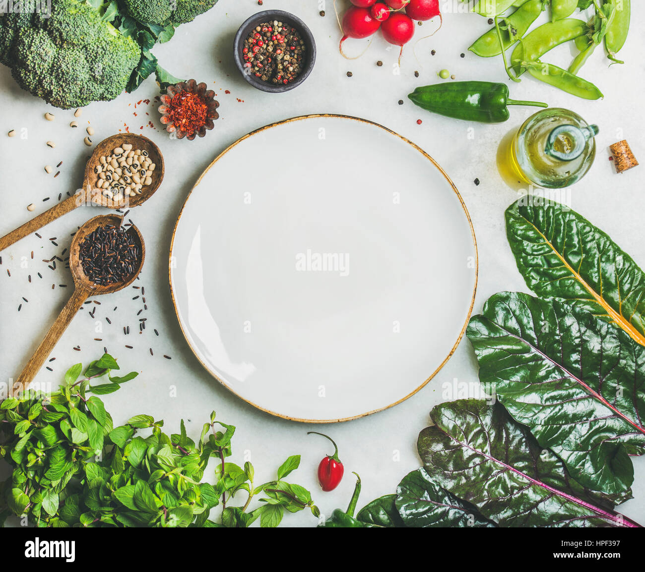 Fresh raw greens, vegetables and grains over light grey marble kitchen countertop, wtite ceramic plate in center, top view, copy space. Healthy, clean Stock Photo