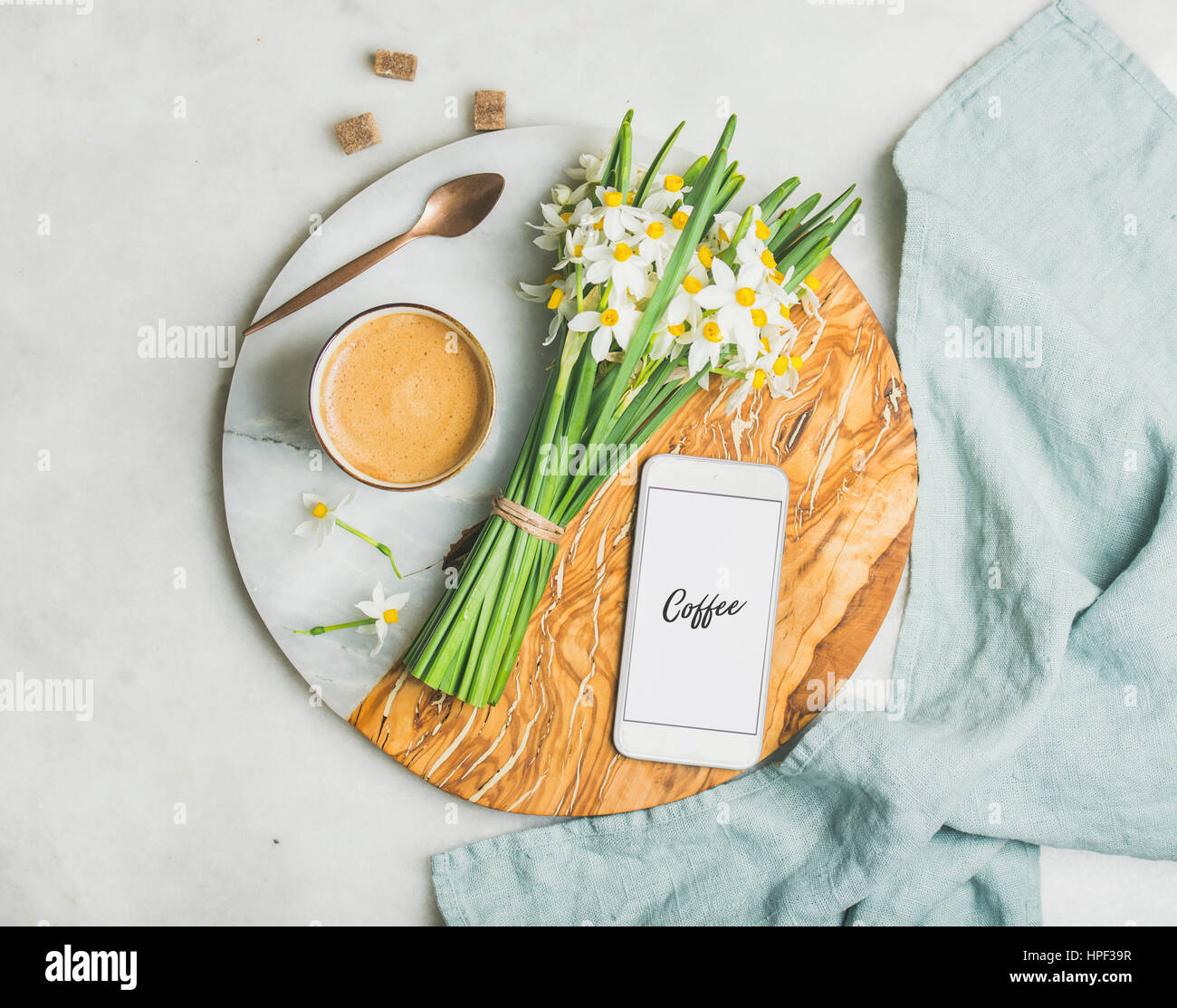 Cup of morning coffee, bucket of spring flowers and mobile phone with text Coffee on serving board over light grey marble background, top view. Mornin Stock Photo