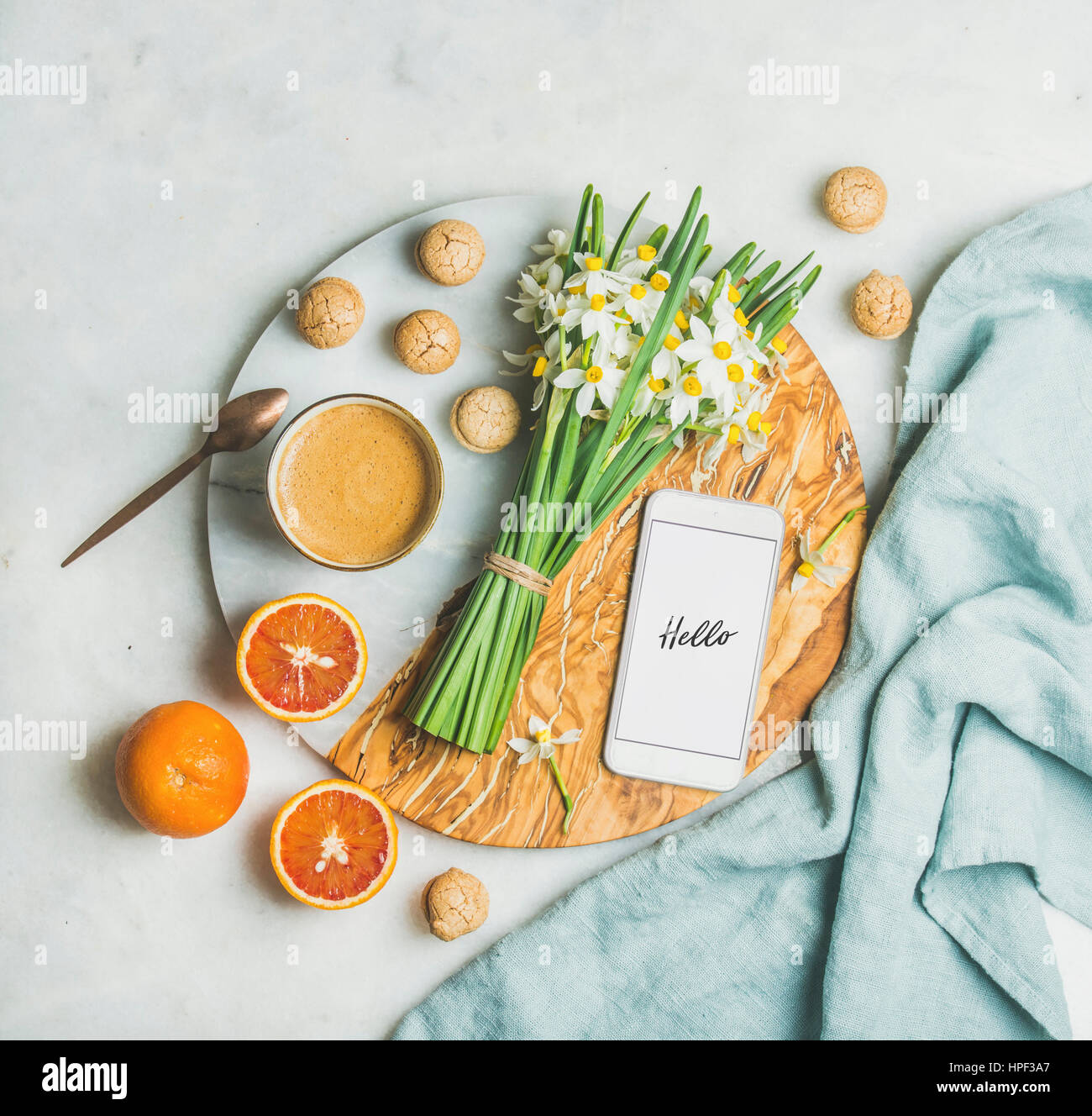 Cup of morning coffee, homemade cookies, red oranges, flowers and mobile phone with text Hello on serving board over light grey marble background, top Stock Photo