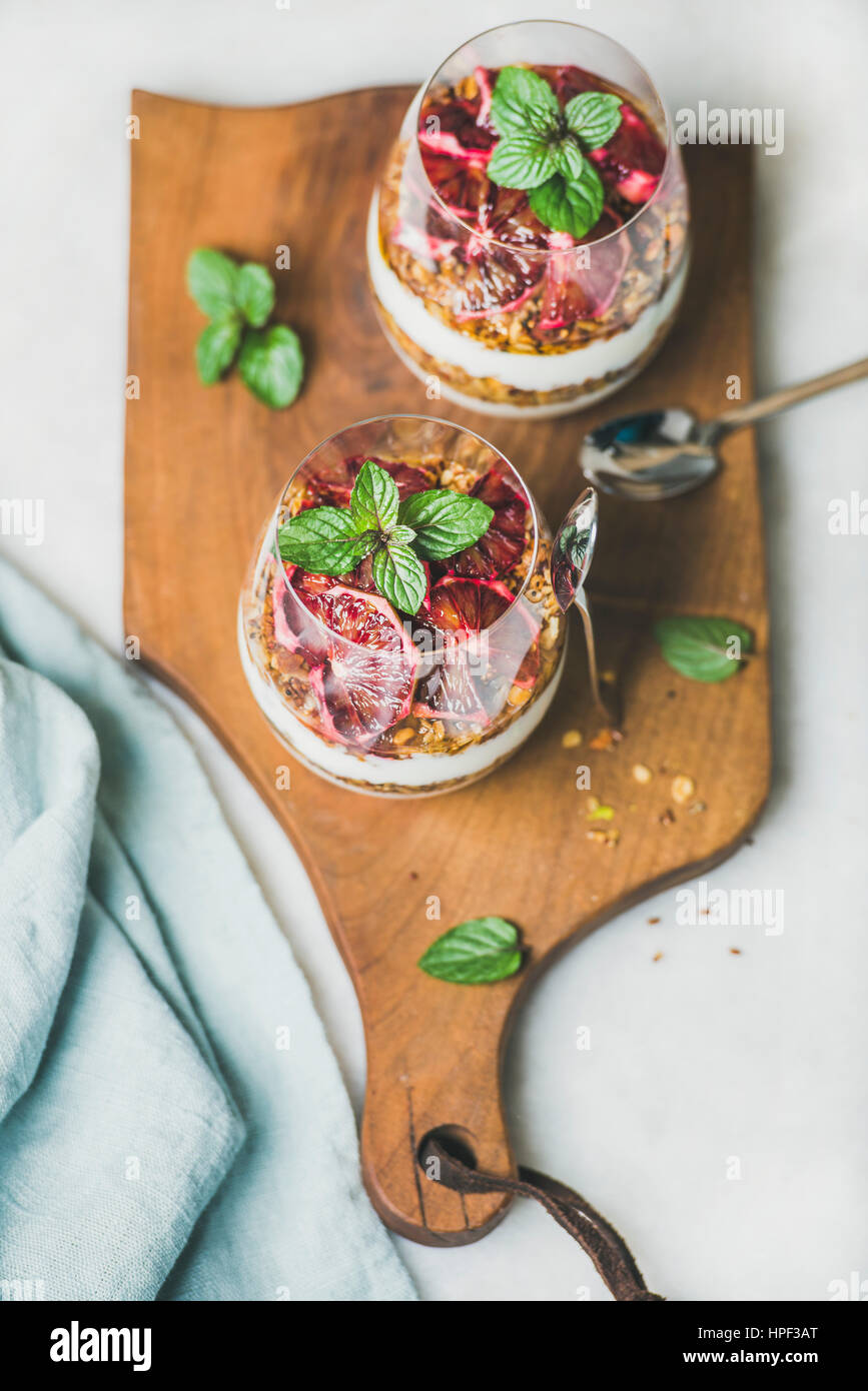 Healthy breakfast. Greek yogurt, granola, blood orange layered parfait in glasses with fresh mint on rustic wooden board over grey marble background. Stock Photo
