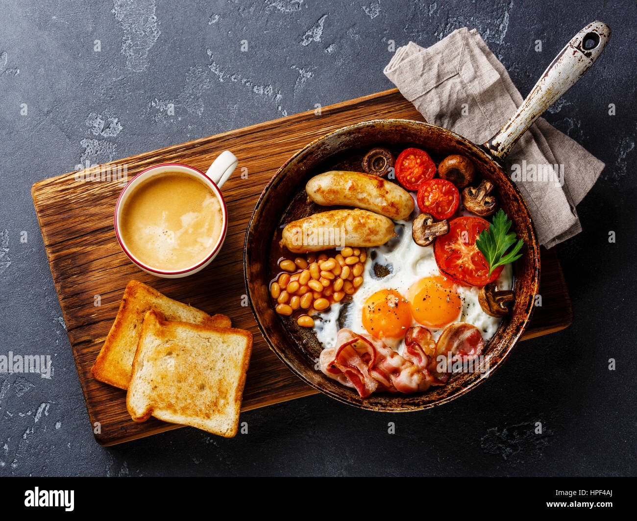 English breakfast in pan with fried eggs, sausages, bacon, beans, toasts and coffee on dark stone background Stock Photo