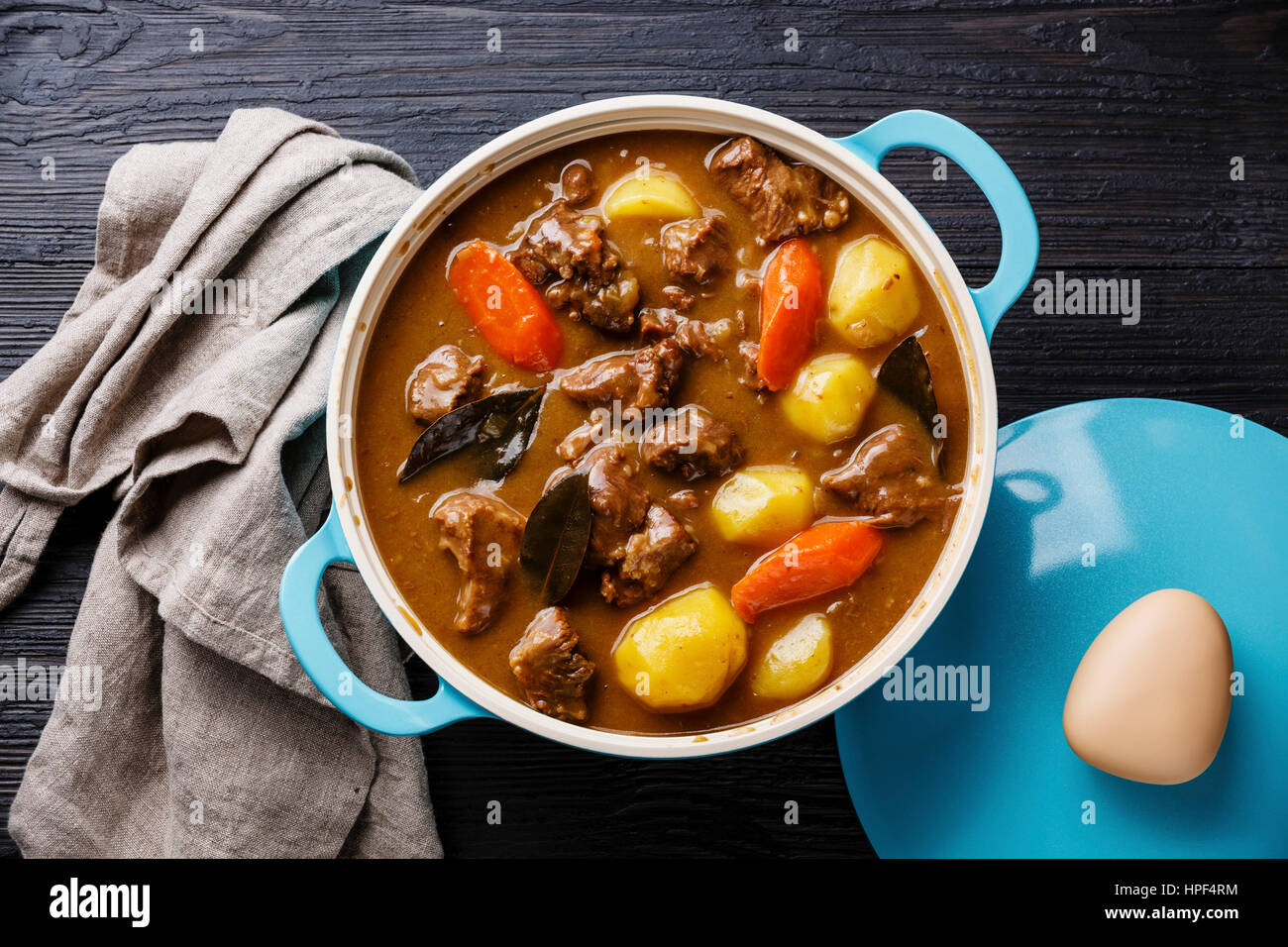 Beef meat stewed with potatoes, carrots and spices in cast iron pan on burned black wooden background Stock Photo