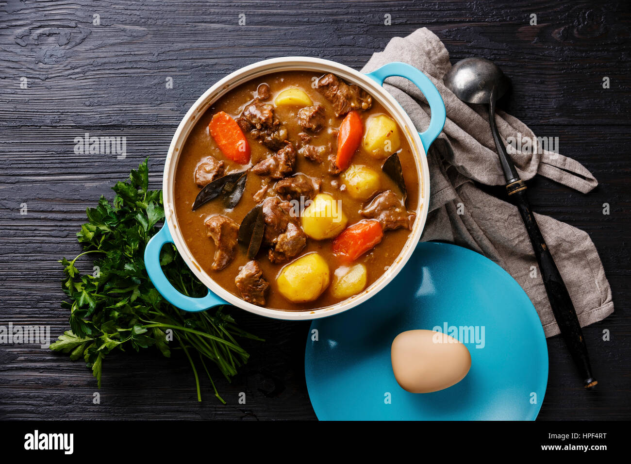 Beef meat stewed with potatoes, carrots and spices in cast iron pan on burned black wooden background Stock Photo