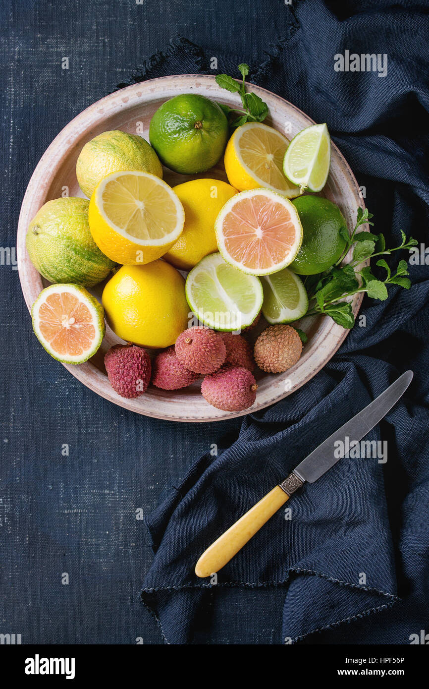 Variety of whole and sliced citrus fruits pink tiger lemon, lemon, lime, mint and exotic lichee on ceramic plate with knife and textile over dark blue Stock Photo