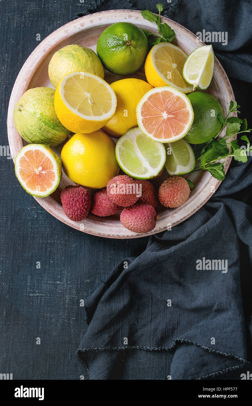 Variety of whole and sliced citrus fruits pink tiger lemon, lemon, lime, mint and exotic lichee on plate with textile over dark blue canvas textured b Stock Photo
