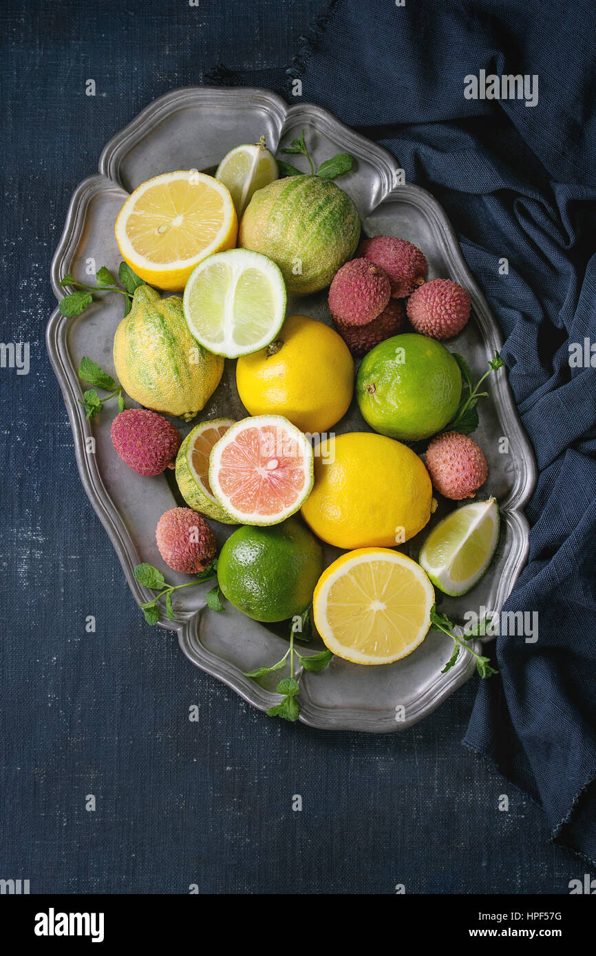 Variety of whole and sliced citrus fruits pink tiger lemon, lemon, lime, mint and exotic lichee on vintage metal plate with textile over dark blue can Stock Photo