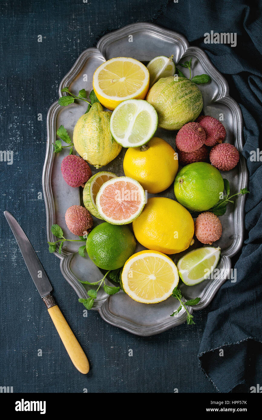 Variety of whole and sliced citrus fruits pink tiger lemon, lemon, lime, mint and exotic lichee on vintage plate with textile and knife over dark blue Stock Photo