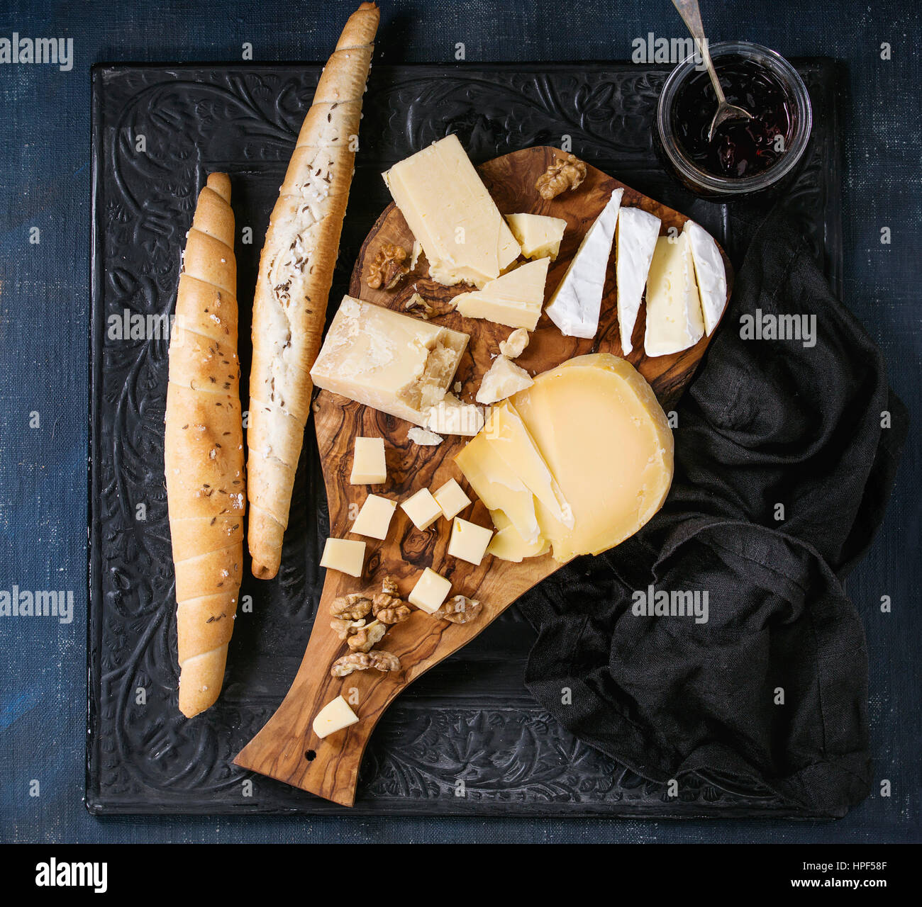 Cheese plate. Assortment of cheese with walnuts, jam and bread on olive wood serving board with textile over black ornate and dark blue canvas as back Stock Photo