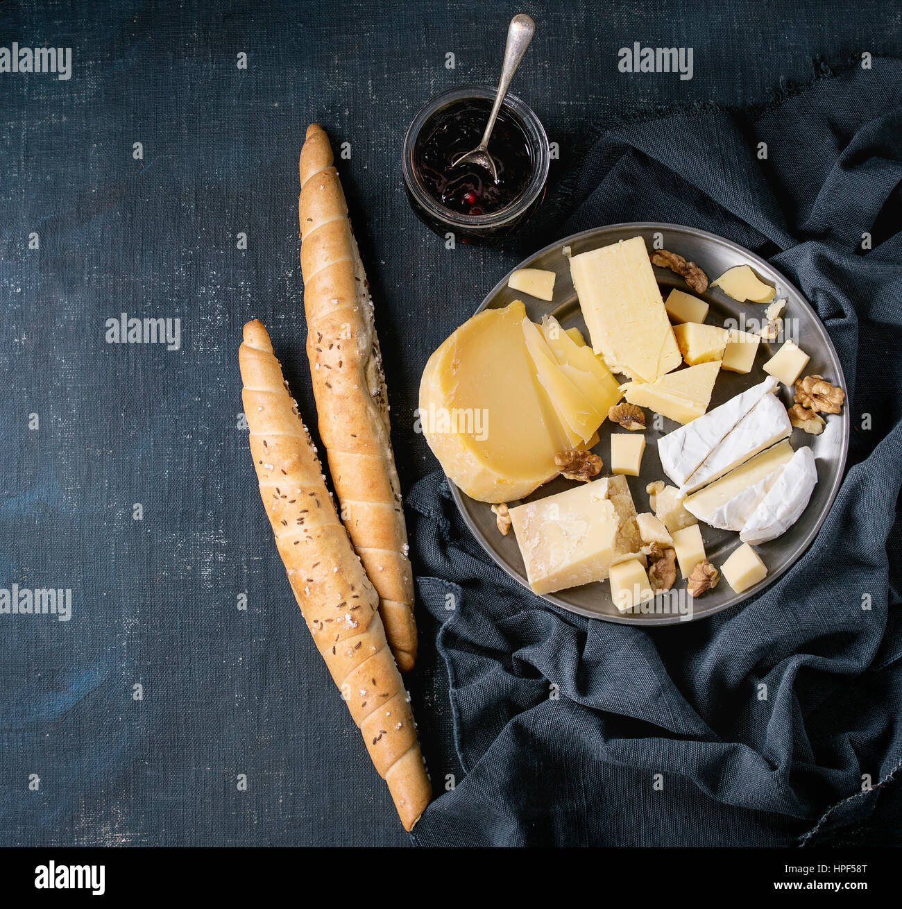 Cheese plate. Assortment variety of cheese with walnuts, jam and bread on vintage metal plate with textile over dark blue canvas as background. Top vi Stock Photo