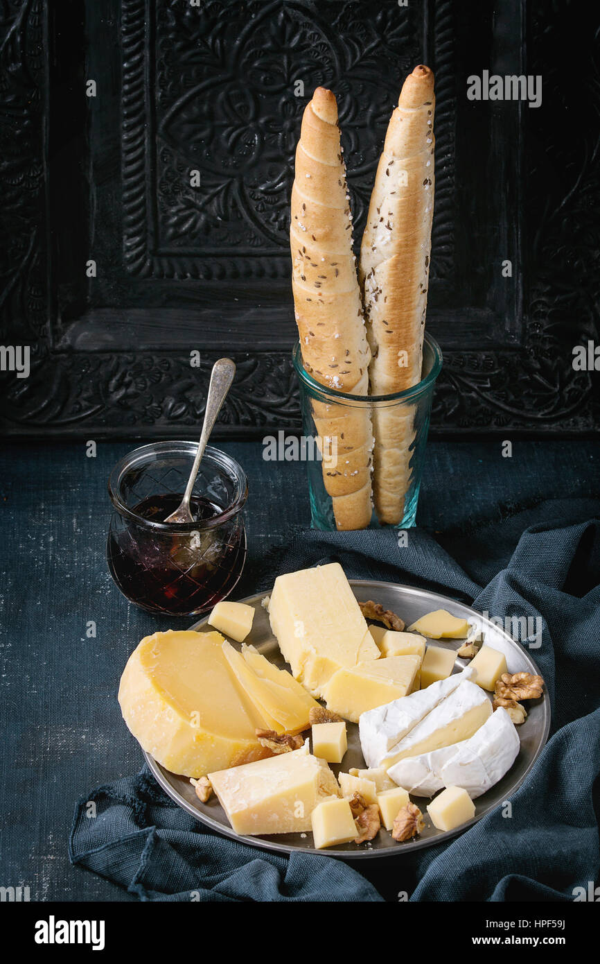 Cheese plate. Assortment variety of cheese with walnuts, jam and bread on vintage metal plate with textile over dark blue canvas with black ornate bac Stock Photo