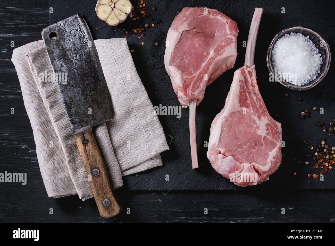 Two raw uncooked Veal tomahawk steak with garlic, salt, seasoning and vintage butcher cleaver on textile napkin on black stone slate board. Top view w Stock Photo