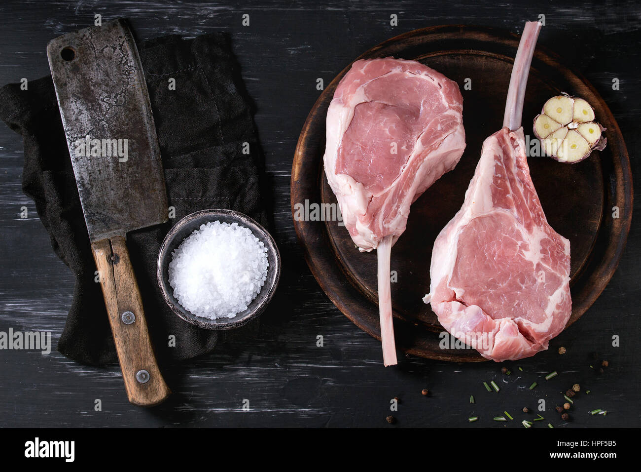 Two raw uncooked Veal tomahawk steak with garlic, salt, seasoning on wooden chopping board and vintage butcher cleaver on textile napkin over black te Stock Photo