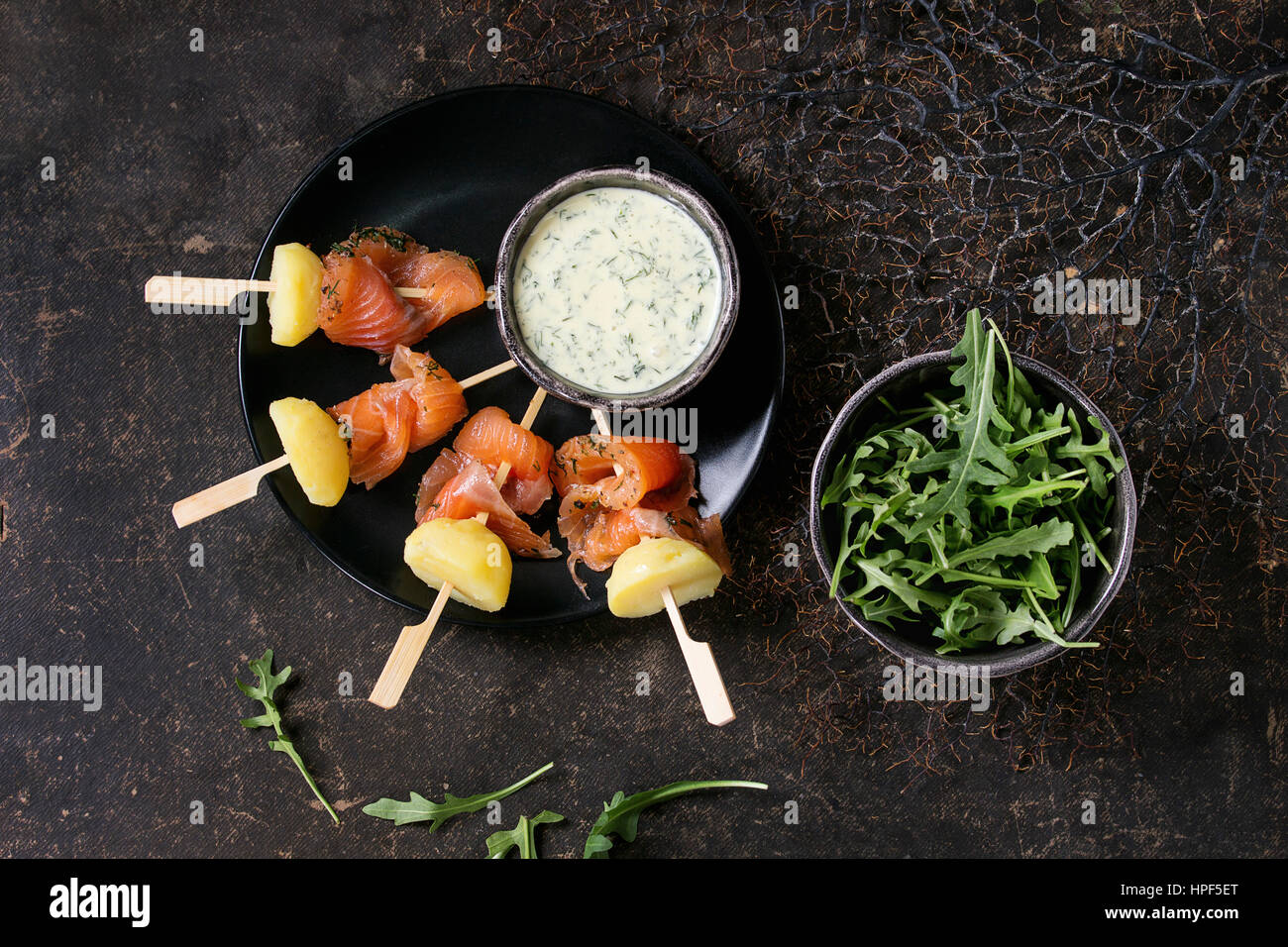 Delicious Appetizer with smoked salted salmon and boiled potatoes on skewers served with creamy dill sauce and arugula on black plate over dark backgr Stock Photo