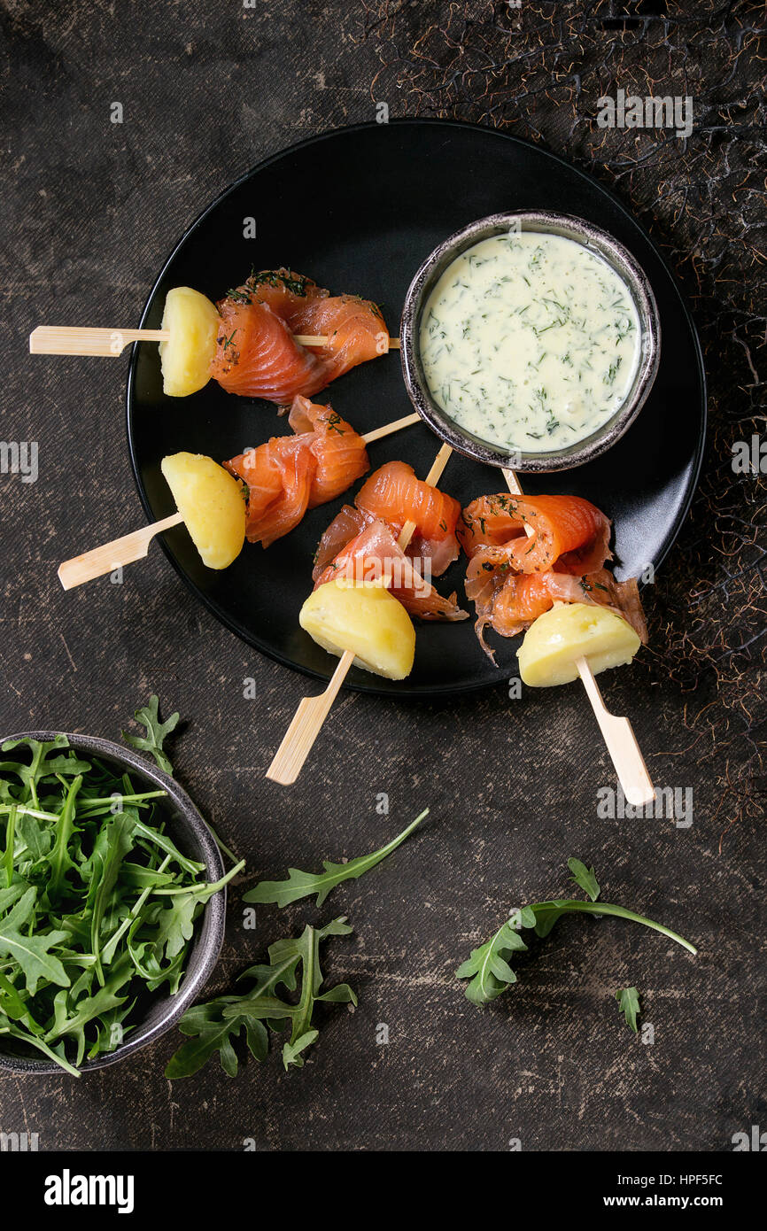 Delicious Appetizer with smoked salted salmon and boiled potatoes on skewers served with creamy dill sauce and arugula on black plate over dark backgr Stock Photo