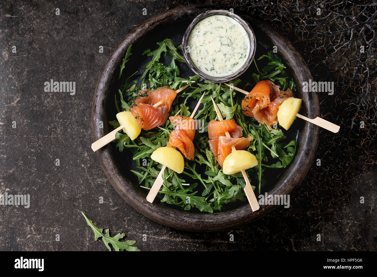Delicious Appetizer with smoked salted salmon and boiled potatoes on skewers served with creamy dill sauce and arugula in stone plate over dark backgr Stock Photo