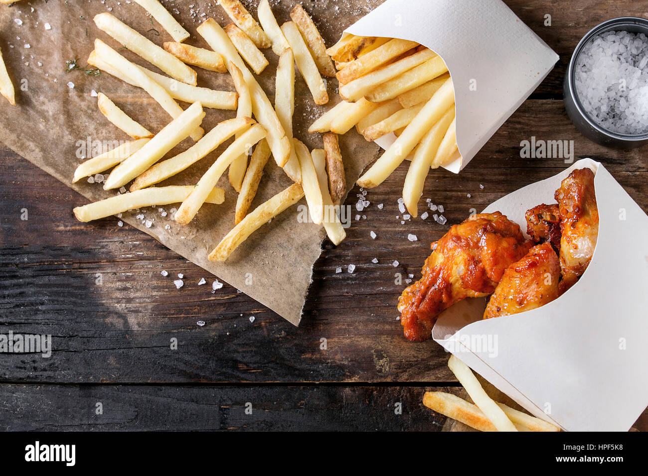 Fast food fried spicy chicken legs, wings and french fries potatoes in lunch boxes with salt and ketchup sauce served on baking paper over old dark wo Stock Photo